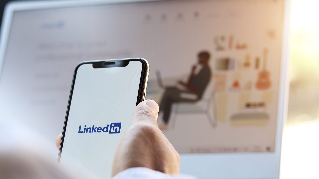 Professional holding cellphone with LinkedIn app on the screen and laptop open with the LinkedIn website open in the background