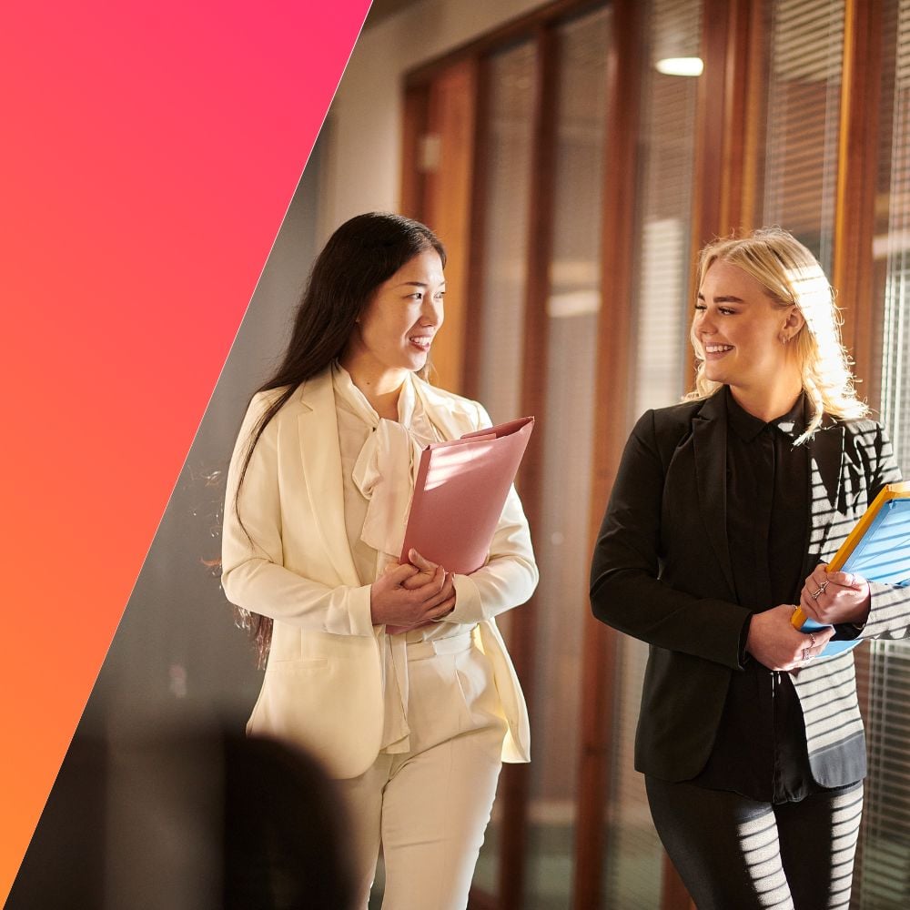 Two professional women, dressed in business attire, walking together in an office hallway, smiling and holding folders, symbolizing collaboration and teamwork in a legal workplace.