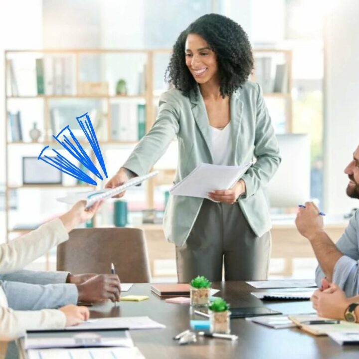 Woman who is a first time manager in an office standing up leading a team meeting handing out paperwork to employees.