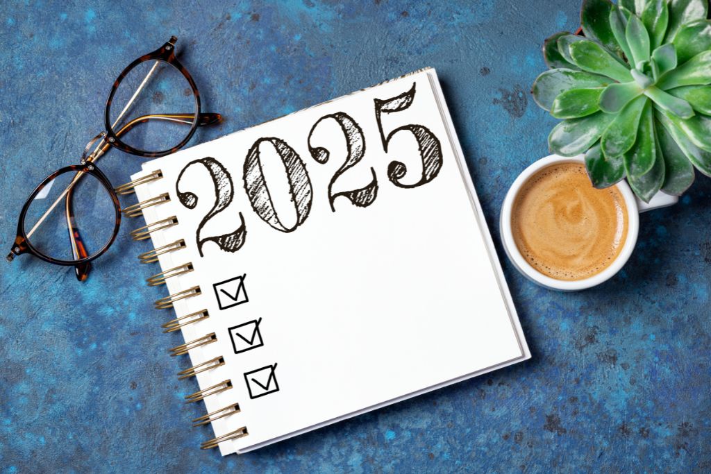 Notebook with '2025' written in bold, sketch-style font and a checklist on the page, placed on a textured blue surface alongside a pair of glasses, a cup of coffee, and a green succulent plant.