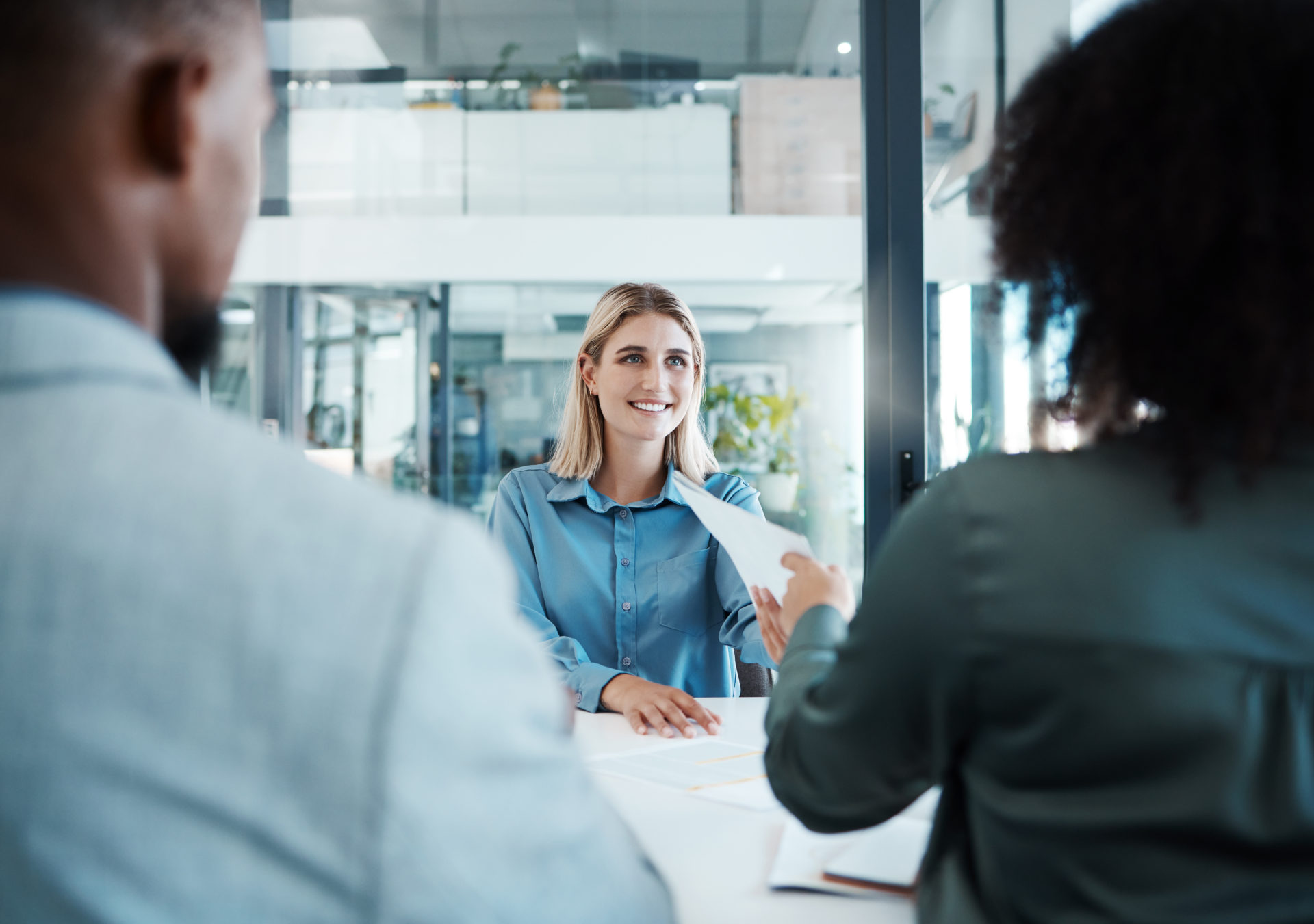 Two hiring managers interviewing a job candidate at a desk in their office. Female job candidate is handing over resume to the female interviewer.