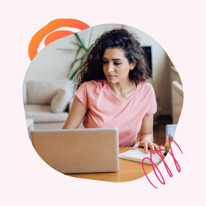 A young woman with curly hair wearing a pink t-shirt is sitting at a wooden table, passively job searching on a laptop with a notebook beside her. She appears focused and engaged, with a comfortable home setting in the background.