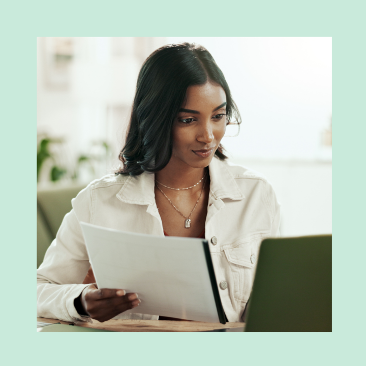 A professional woman in a light-colored jacket holding summer job cover letter while looking at her laptop screen. She appears focused, working in a bright and modern setting.