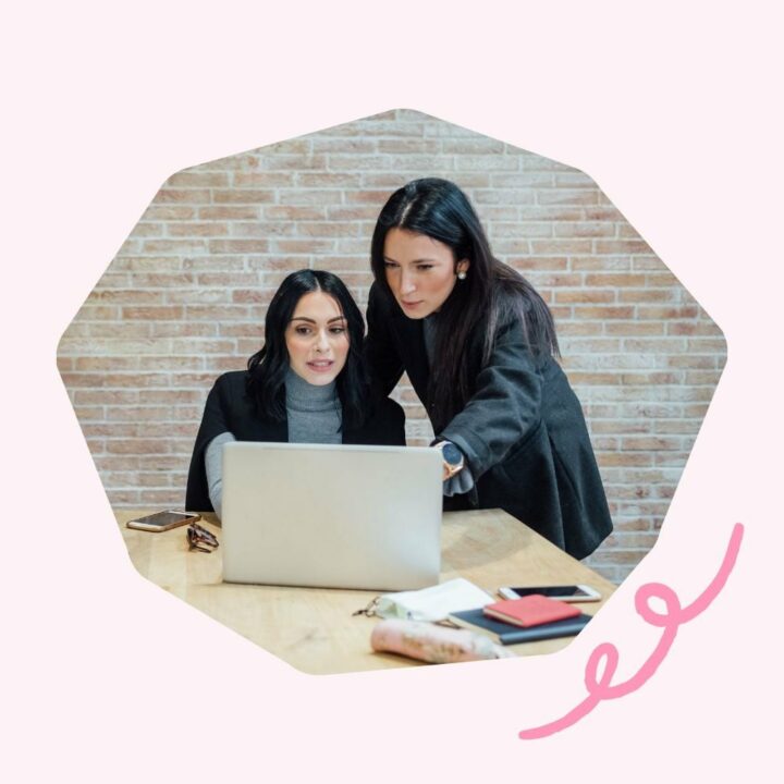 Two women with dark hair are working together at a desk, looking intently at a laptop screen. One woman is sitting while the other is standing, pointing at the screen.