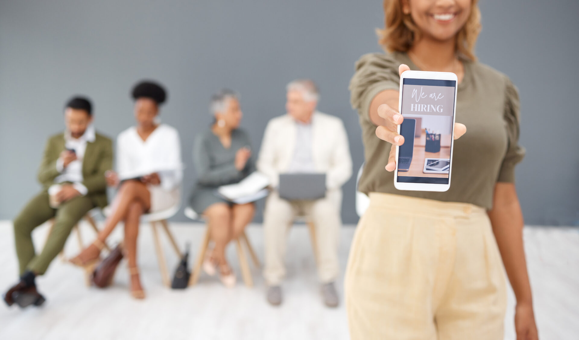 Woman holding smartphone with picture that says "we are hiring," and people waiting in the background with resumes for recruiting event