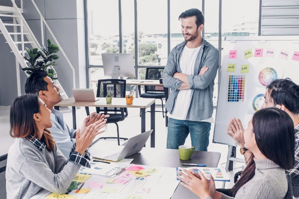 Manager mapping out an employer branding strategy on white board with sticky notes and four employees discussing and clapping sitting around a table