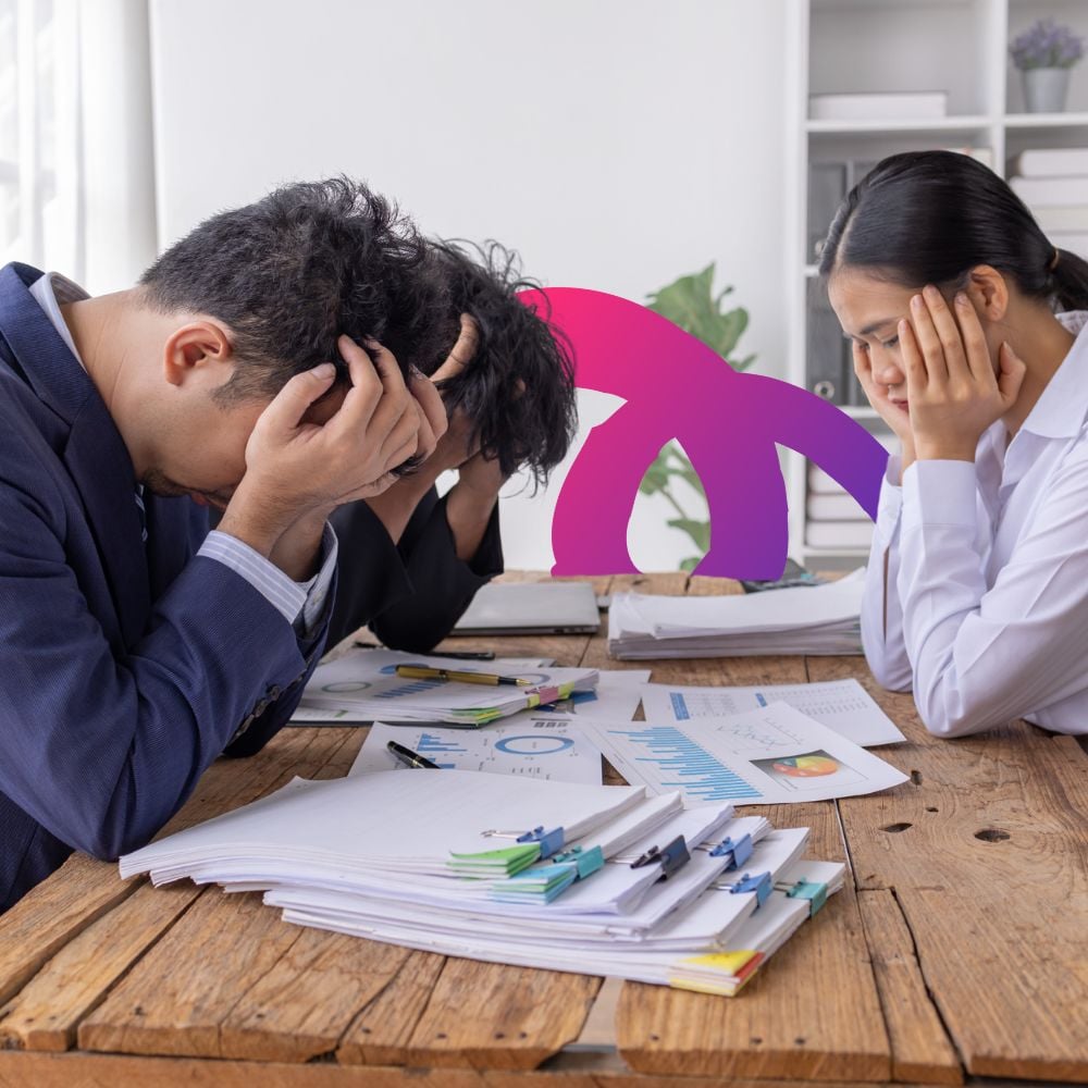 Three professionally dressed employees looking stressed and rubbing their temples sitting around a desk with piles of paperwork in front of them. Bad company culture concept.