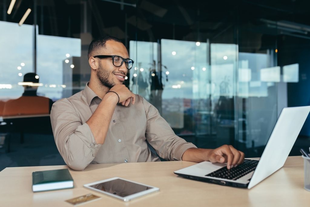 Recruiter wearing glasses resting his hand on his face at his desk on a laptop tracking hiring KPIs