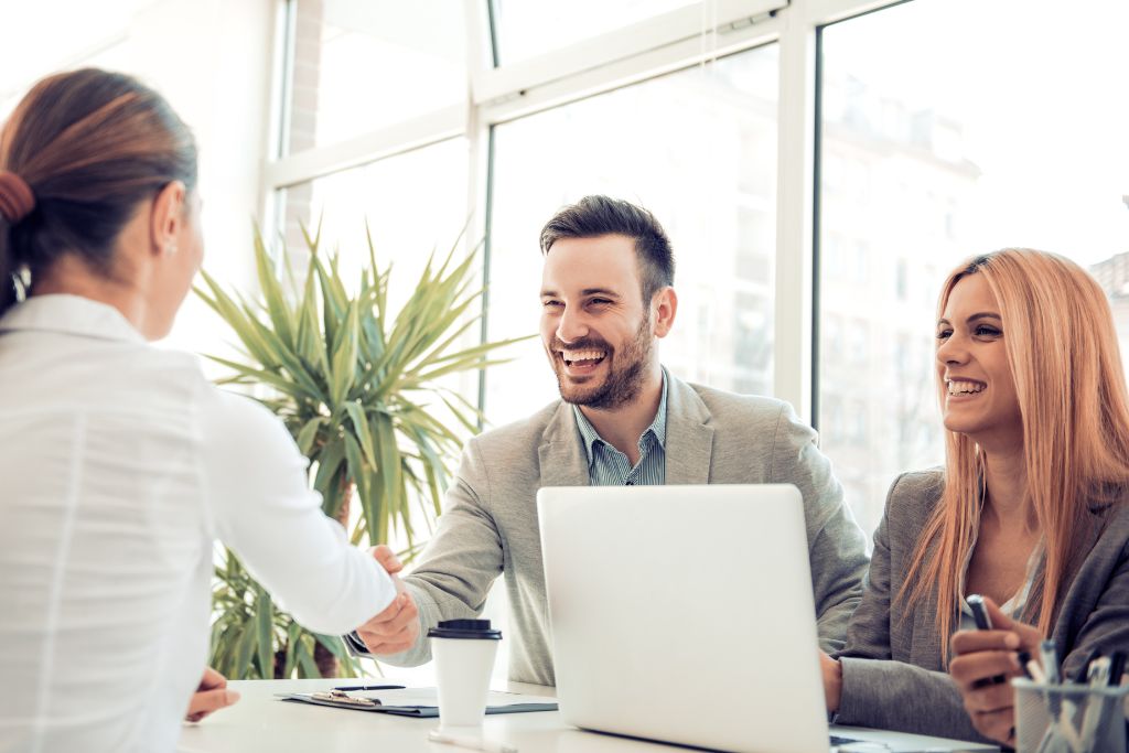 Two good interviewers dressed professional sitting at a desk behind a white laptop shaking the hand of a female job candidate in a white blouse