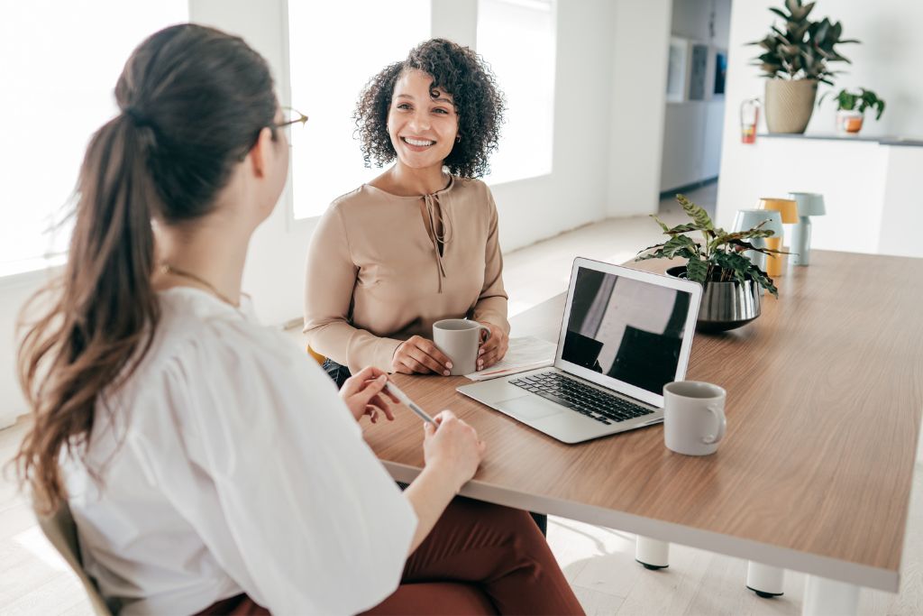 Female hiring manager topgrading interviewing a female job candidate sitting at a modern desk with her laptop open and a cup of coffee right in front.
