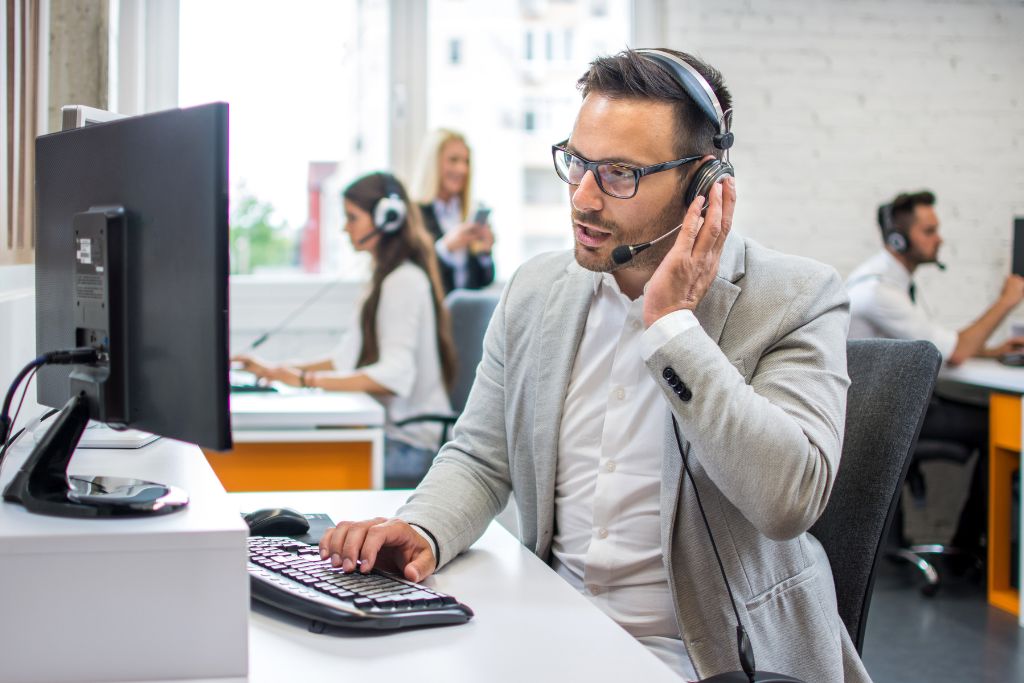 Call center employee in a gray suit sitting at a desk in front of a desktop computer talking into a headset