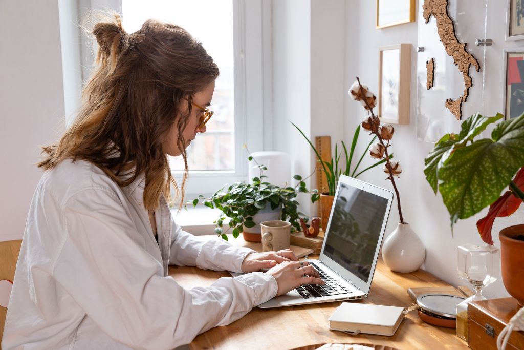 Female recruiter wearing a white blouse and red glasses typing an interview invitation email on her laptop sitting at her home desk next to a few plants