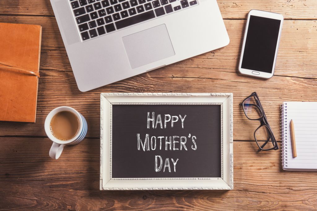 View of a desk with laptop open, cell phone, notebook, cup of coffee, and a message board that says happy mother's day