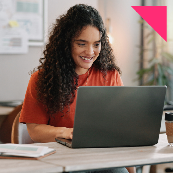 A smiling woman with curly hair wearing an orange shirt is working on her laptop in a modern, well-lit workspace. An open notebook and coffee cup sit on the table beside her as she types.