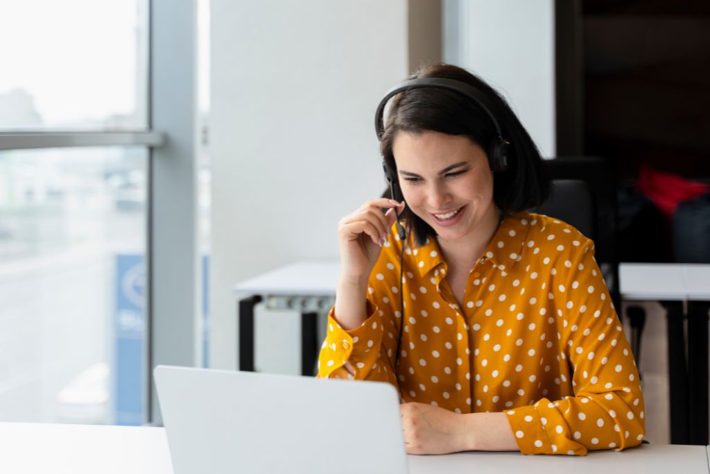 Smiling recruiter woman wearing a yellow polka dot shirt and talking into a headset, working on a laptop in a modern office setting.