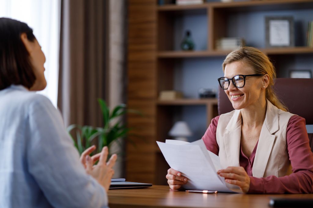 Female hiring manager for a small business smiling wearing black glasses and a beige blazer is sitting in a modern office across from job candidate during an interview