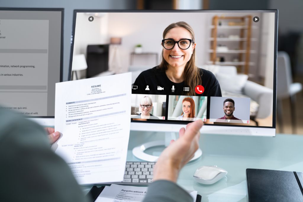 A close-up of a video interview setup showing the interviewer holding a resume and the smiling female candidate on the computer screen.