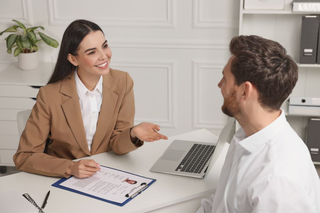 A professional job interview setting featuring a talent acquisition woman in a beige blazer conducting an interview with a male candidate. The woman is smiling and engaged, holding a clipboard with a resume visible on top. The man, seated across from her, appears attentive and is dressed in a white shirt. A laptop is open in front of the woman on a white desk, which also has a pen and notebook. The background includes white paneled walls and office shelves, creating a formal and organized atmosphere.