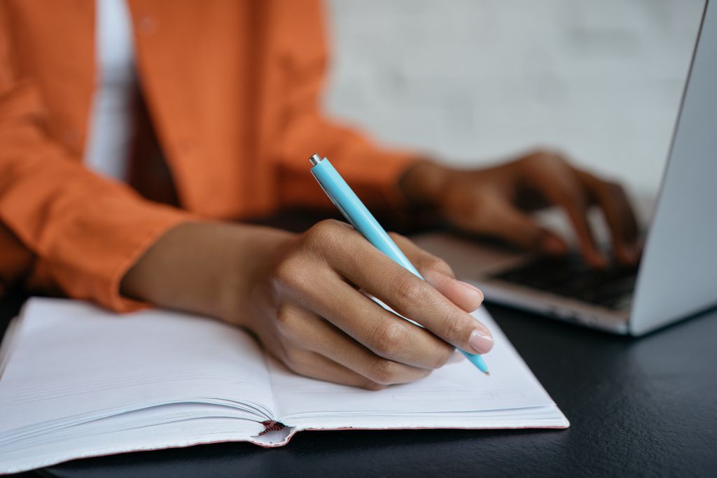 A hiring manager in an orange jacket writing in a notebook with a light blue pen, while using a laptop, taking notes during an interview