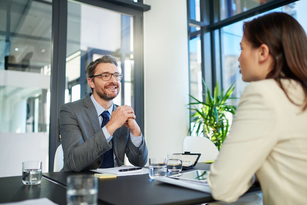 A professional man in a suit conducts a competency-based interview with a woman. Both are seated across a table in a modern office with large windows, natural light, and a few plants in the background. The man is smiling and listening attentively while the woman responds.