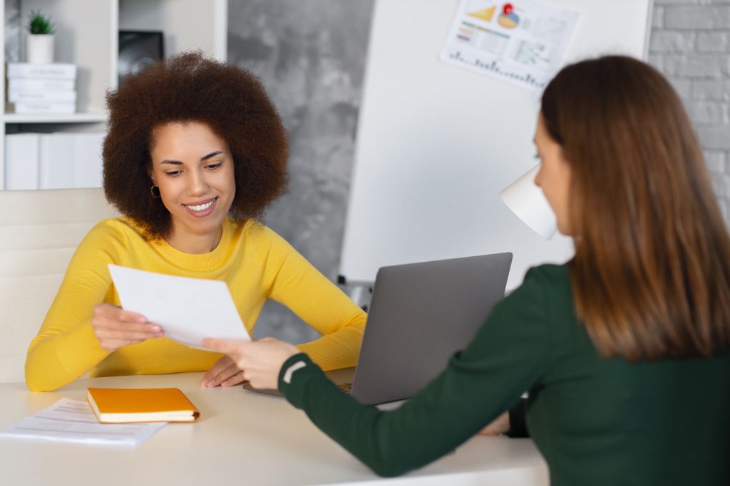 Two women are engaged in a professional setting. One woman, with curly hair and wearing a yellow top, is smiling while reviewing a document handed to her by the other woman, who has brown hair and is wearing a green top. The second woman is seated across from her, holding a paper, with a laptop and some documents on the table between them. The setting appears to be an office environment, with charts on a whiteboard in the background.