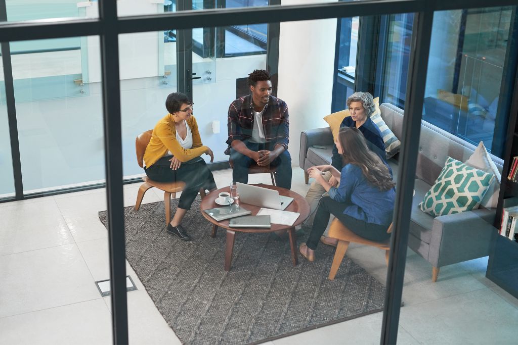 A group of four people is seated in a modern office lounge, engaged in a casual meeting about partnering with staffing agency. Two women and two men sit around a small round table with laptops and coffee cups. One woman in a mustard-yellow sweater leans forward, while another gestures as she speaks. The others listen attentively, creating a collaborative and relaxed atmosphere with glass walls and cozy seating.