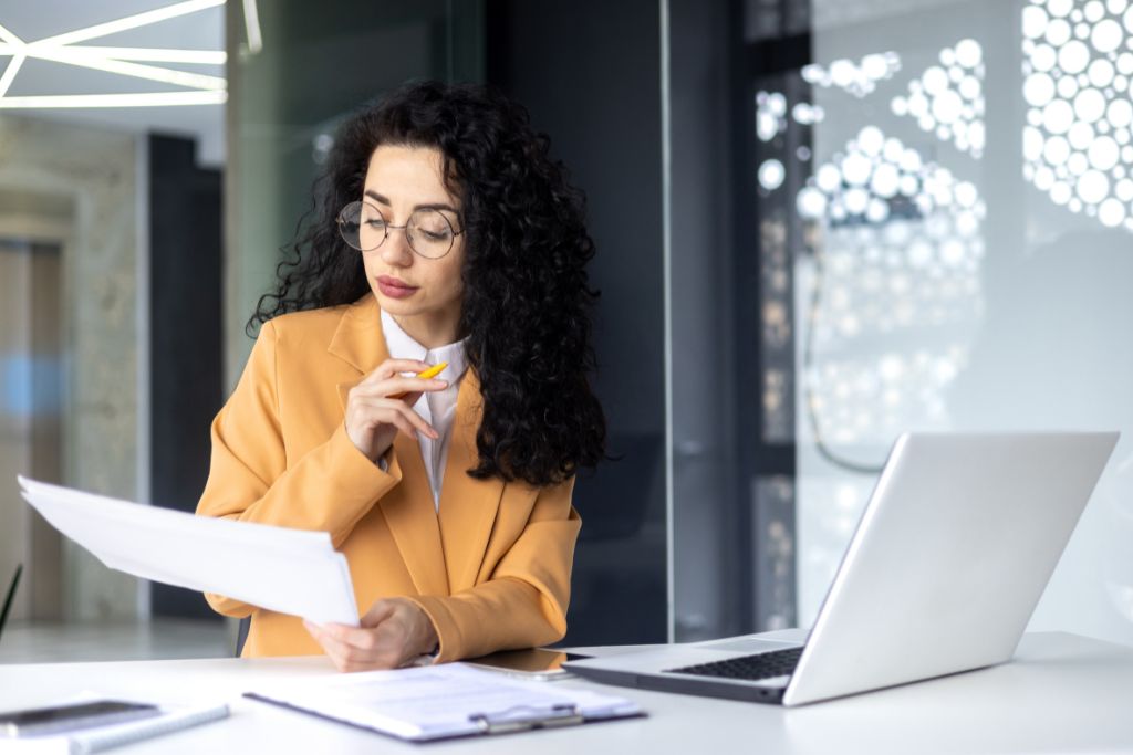 A professional woman with curly hair, wearing glasses and a mustard-colored blazer, thoughtfully reviews a hiring needs document at her desk. She holds a pen to her lips while analyzing the paperwork, with a laptop and clipboard nearby in a modern office setting.