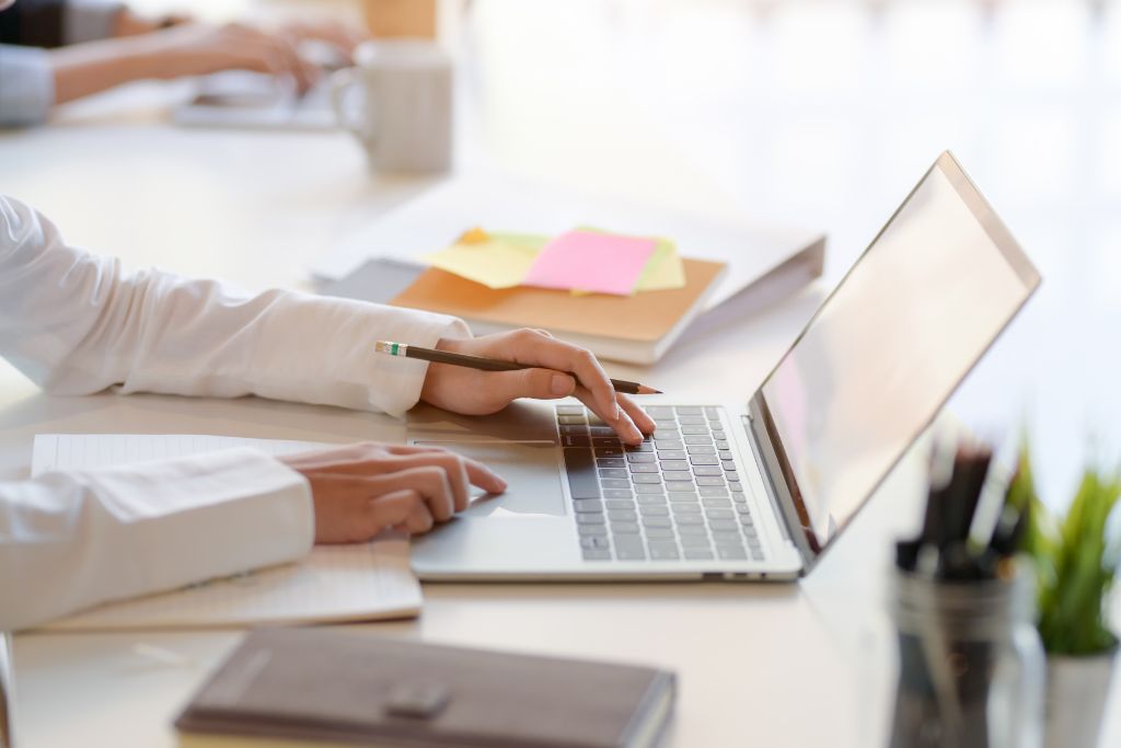 A person in a white shirt creating a job posting on a laptop at a clean, minimal desk. The individual is typing with one hand while holding a pencil in the other hand. Notebooks, a cup, and colorful sticky notes are visible in the background, giving the scene a professional and organized feel.