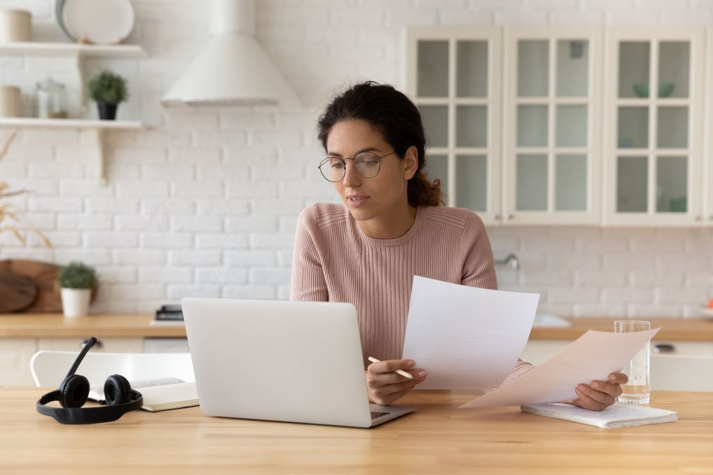 A woman wearing glasses, sitting at a kitchen table, reviewing job applications while working on a laptop, with headphones and a notebook placed nearby.