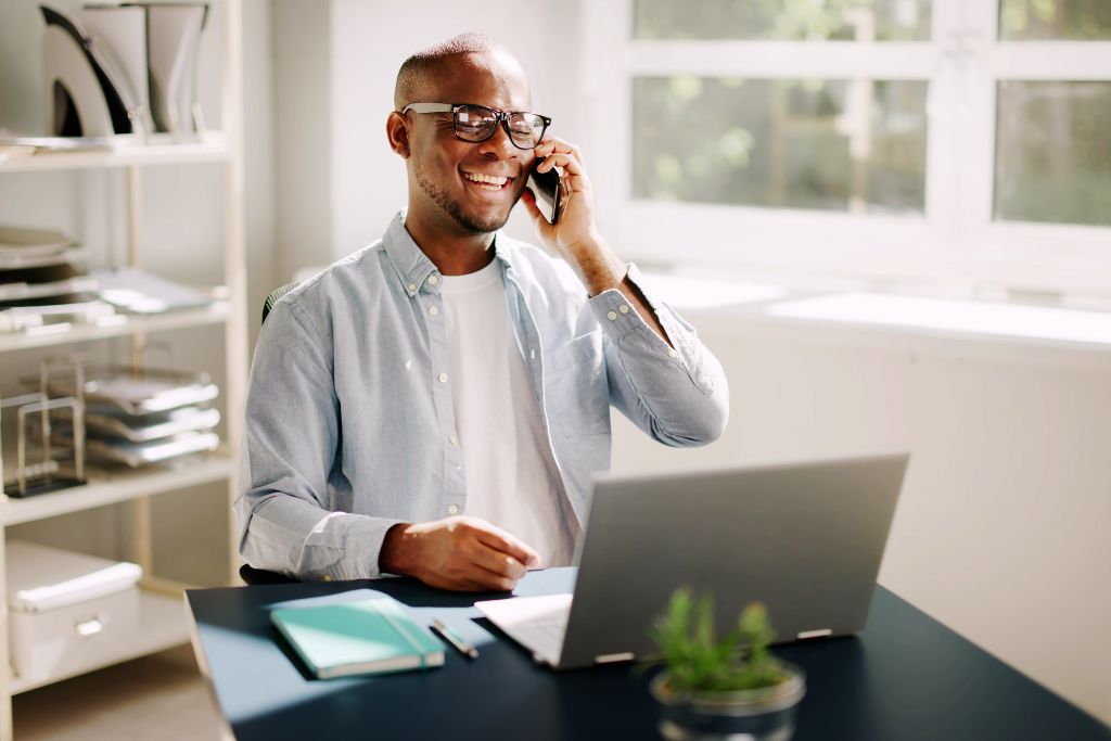 Smiling professional male recruiter wearing glasses and a light blue shirt, sitting at a desk with a laptop, on a call with a jo seeker in a bright office environment.