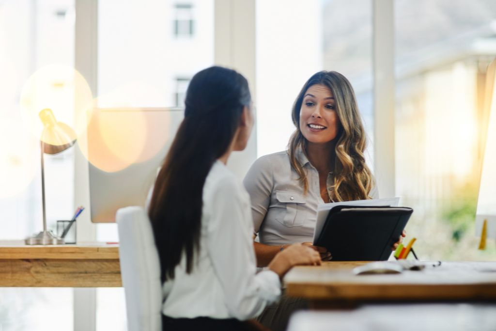 Two professionals engaged in a conversation during a job interview, with a hiring manager smiling and holding a clipboard in a bright, modern office setting