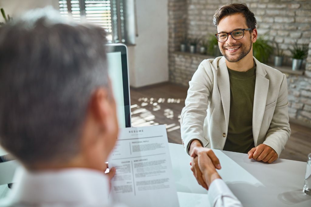 Candidate and interviewer shaking hands across a desk during a job interview, with a resume visible in the foreground, symbolizing a positive and professional interaction.