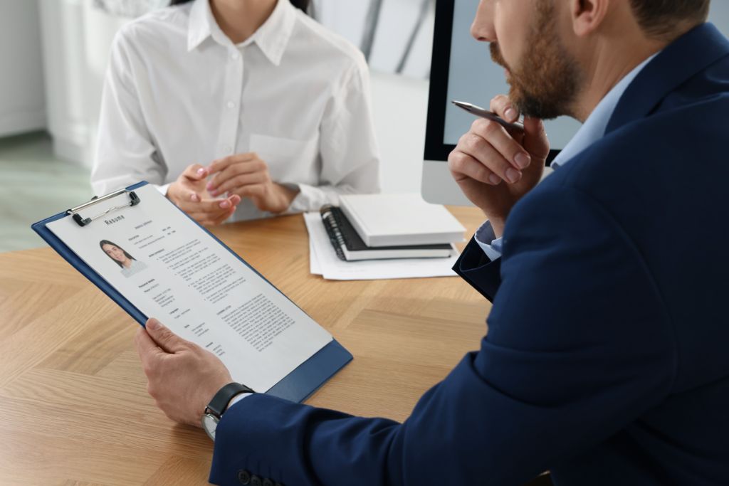 A professional job interview scene showing a recruiter in a suit reviewing a candidate's resume on a clipboard while the candidate, wearing a white shirt, sits across the table, with a computer and notebook in the background.