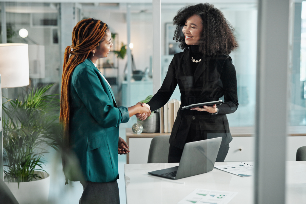 Two professional women in an office shaking hands during an accounting interview. One holds a tablet, while a laptop and financial documents are visible on the desk.