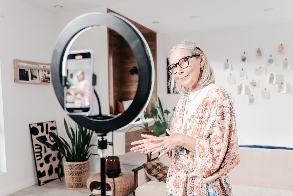 An older woman professional recruiter with gray hair and glasses filming job promotion content using a smartphone and ring light in a modern, stylishly decorated living room. She is wearing a floral-patterned kimono and gesturing as she speaks to the camera.